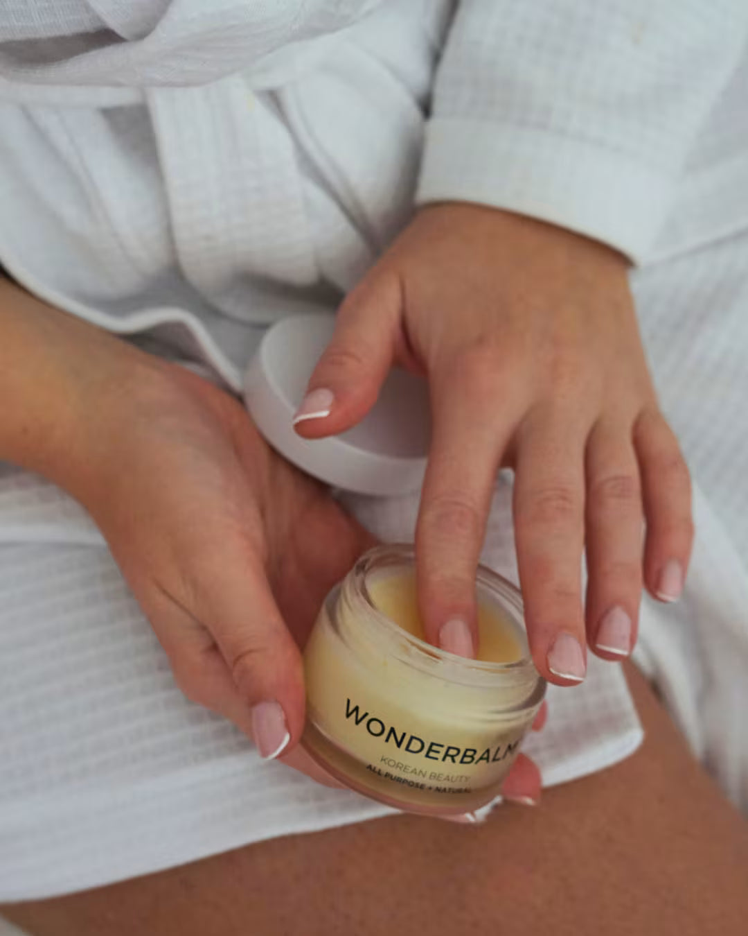 Person holding and using a jar of WONDERBALM with a white robe on.